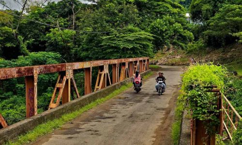 An iron bridge in a lush jungle landscape with two men on motorcycles on it