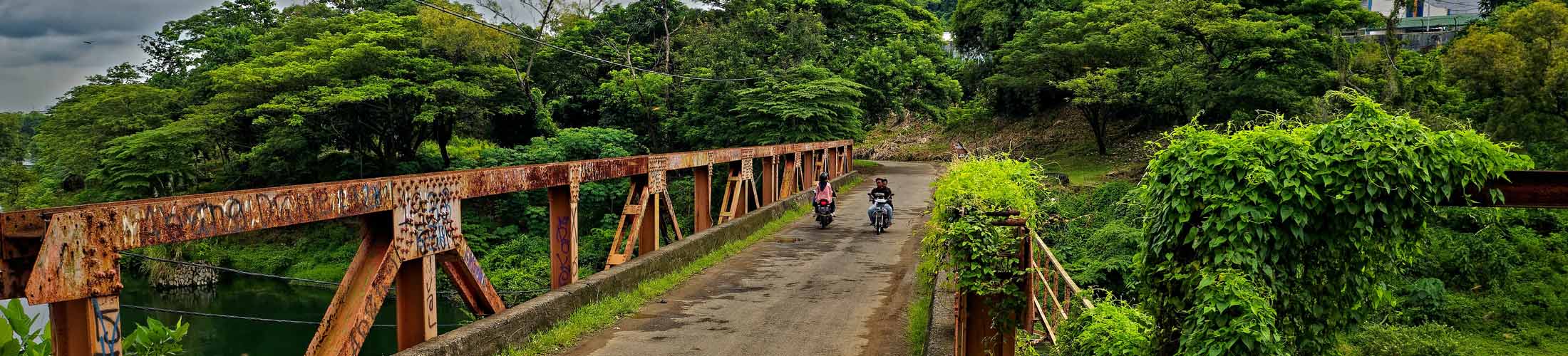 An iron bridge in a lush jungle landscape with two men on motorcycles on it