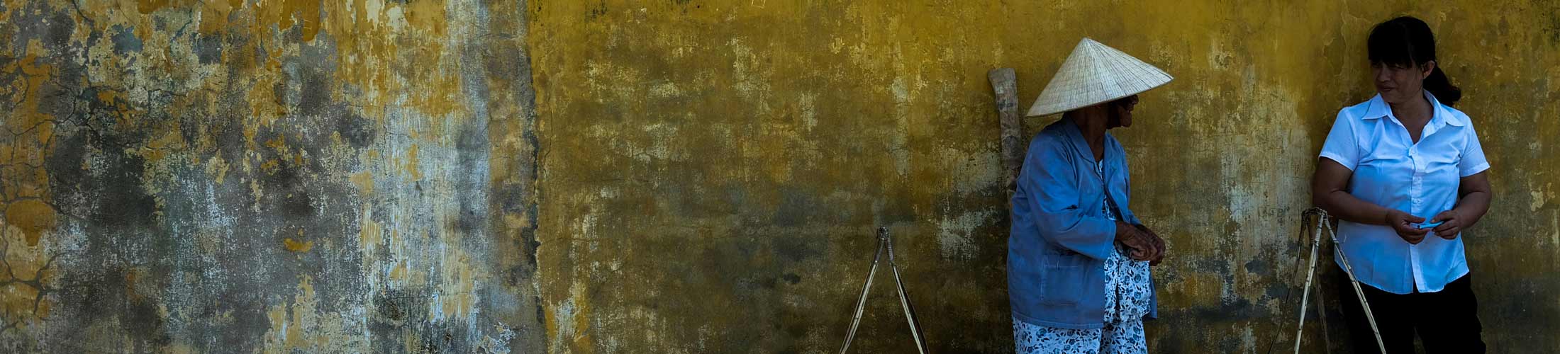 An elderly woman and a younger woman engaged in conversation against a textured yellow wall